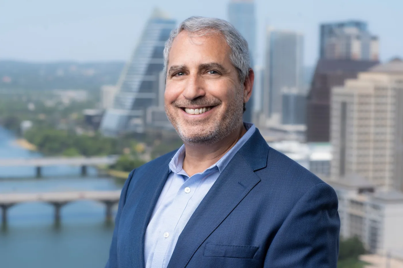 Rich Edison smiling in a navy blazer, with a river, bridges and downtown towers behind him