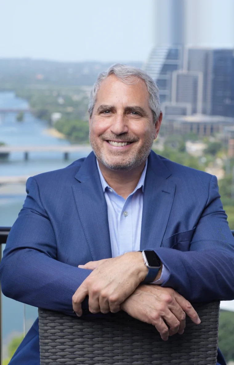 Rich Edison in a navy blazer, arms folded over a chair, with a river and city skyline behind him